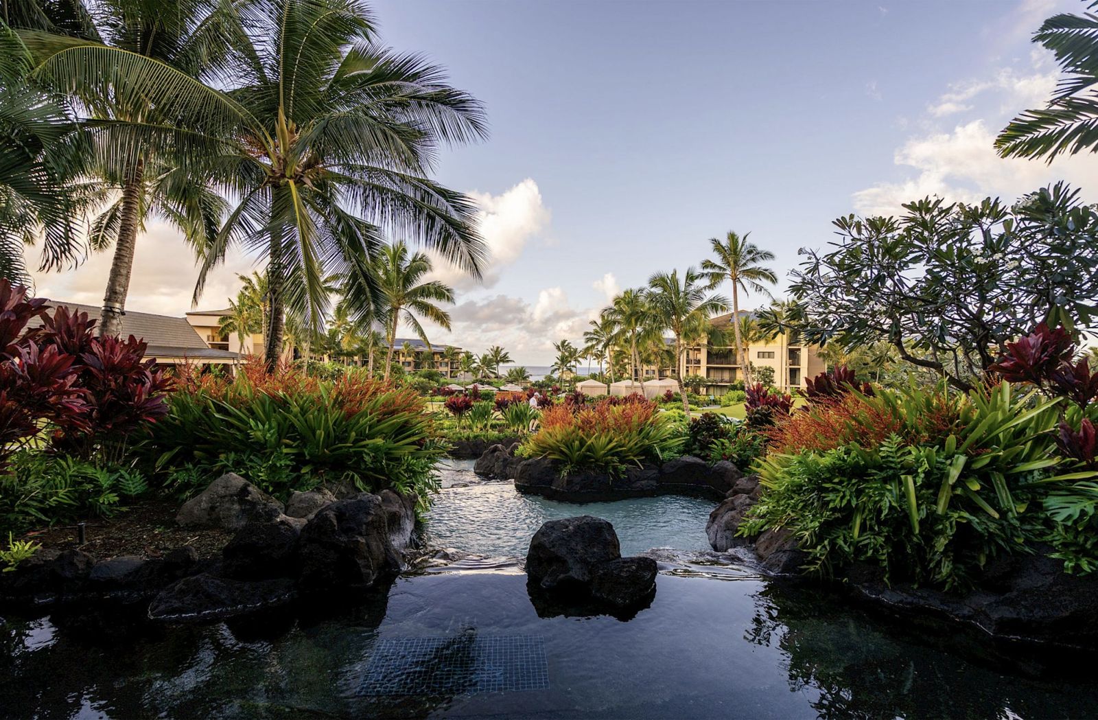 Lagoon-style pool winding between lava rock and tropical gardens at Koloa Landing Resort, palm trees against a soft evening sky